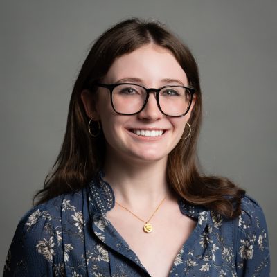 headshot of a women with brown hair and glasses in front of a grey background